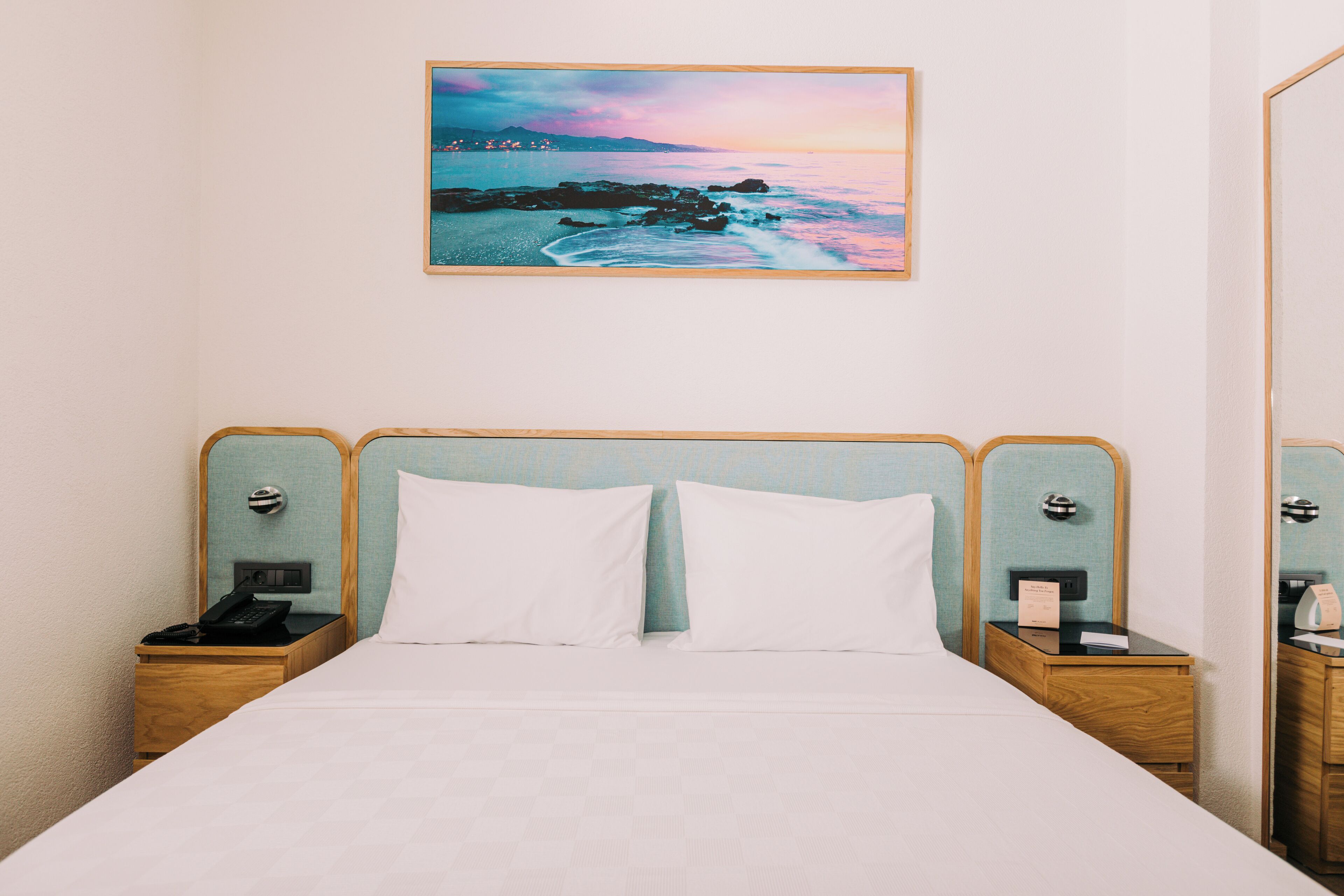 Habitación de hotel con una cama, dos mesitas de noche, un búcaro y un cuadro de una escena costera.