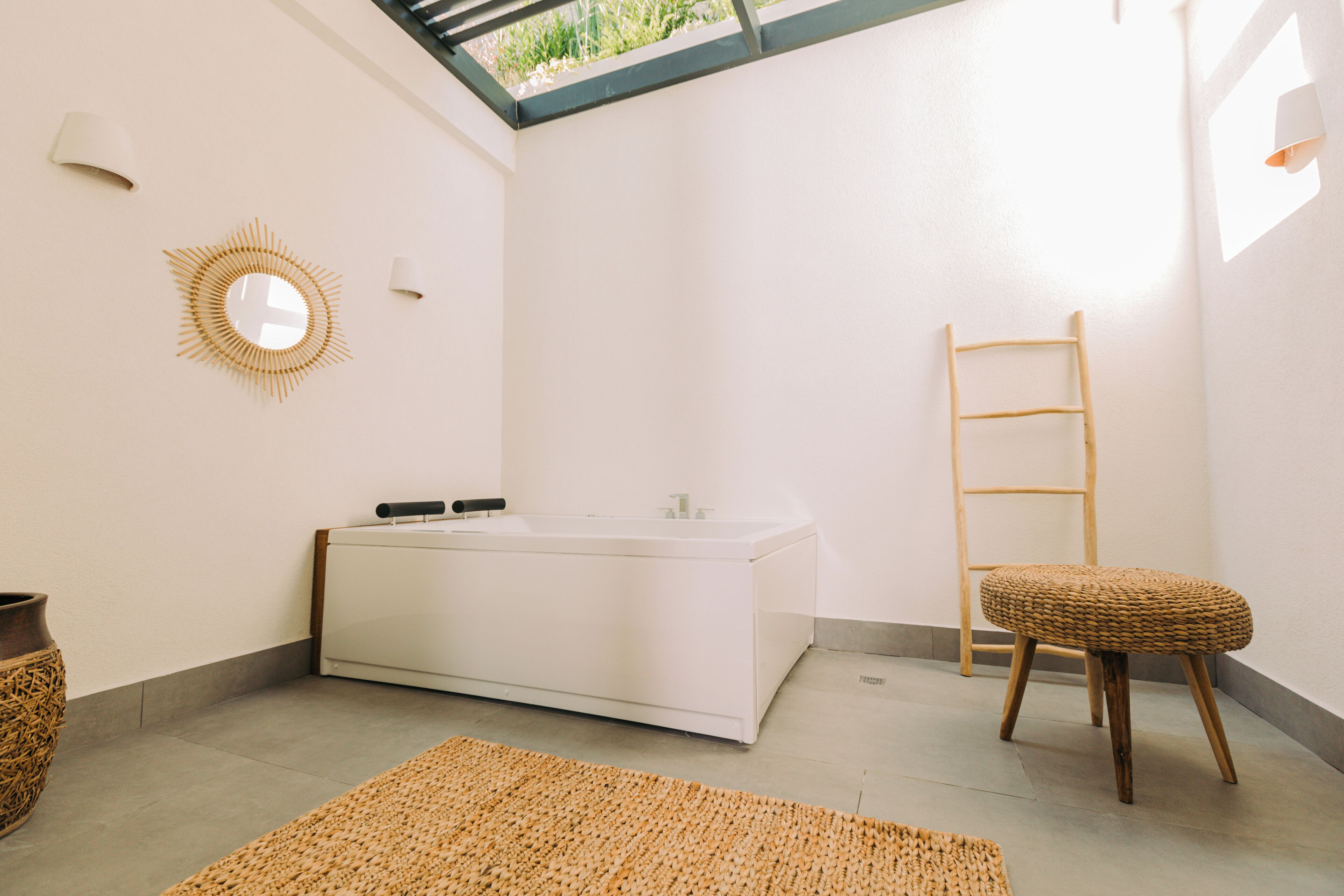A bathroom with a white bathtub, a wooden stool, a woven rug, and a sunburst mirror on the wall.