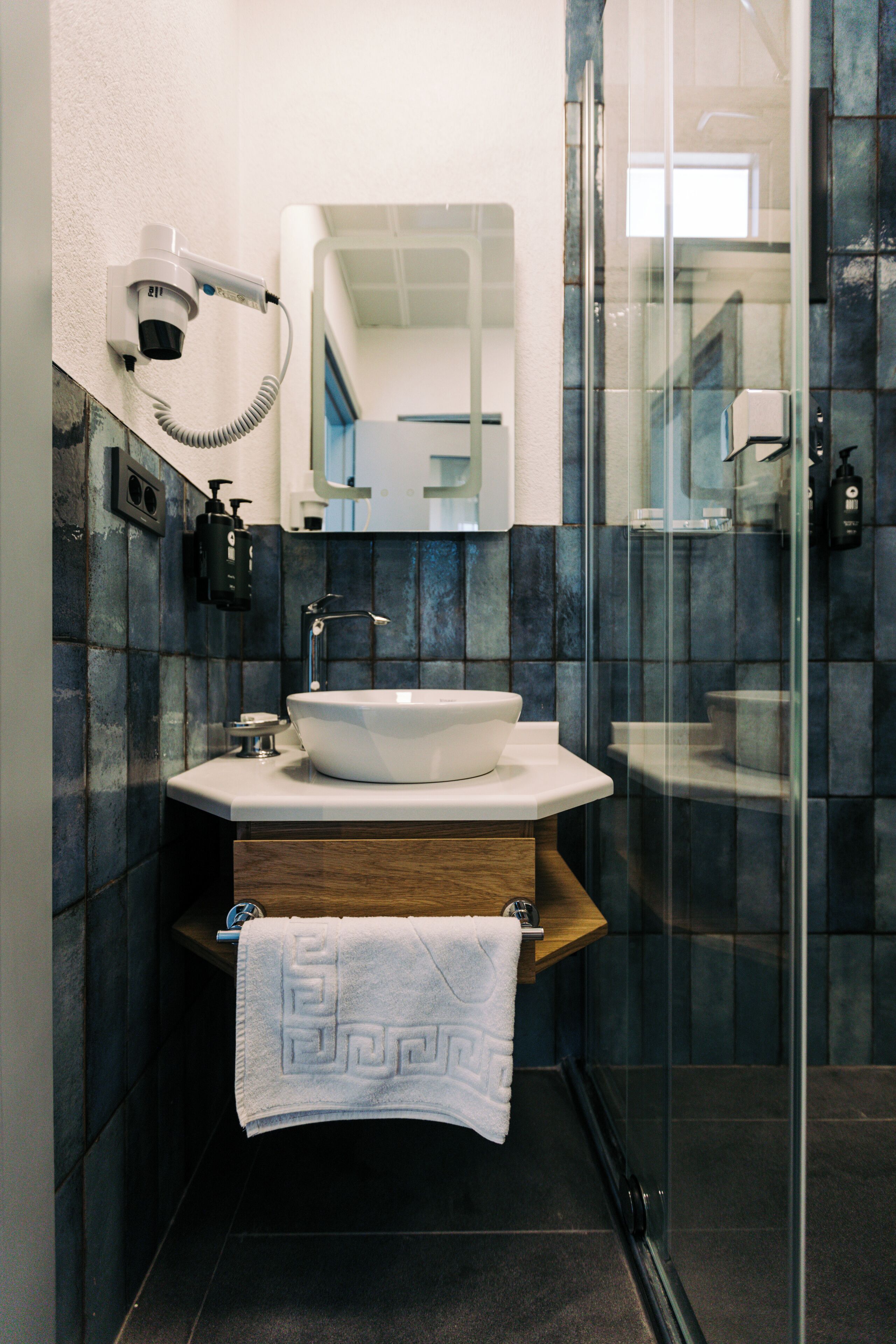 A modern bathroom with a white sink, a glass shower enclosure, and a towel hanging on a wooden shelf.