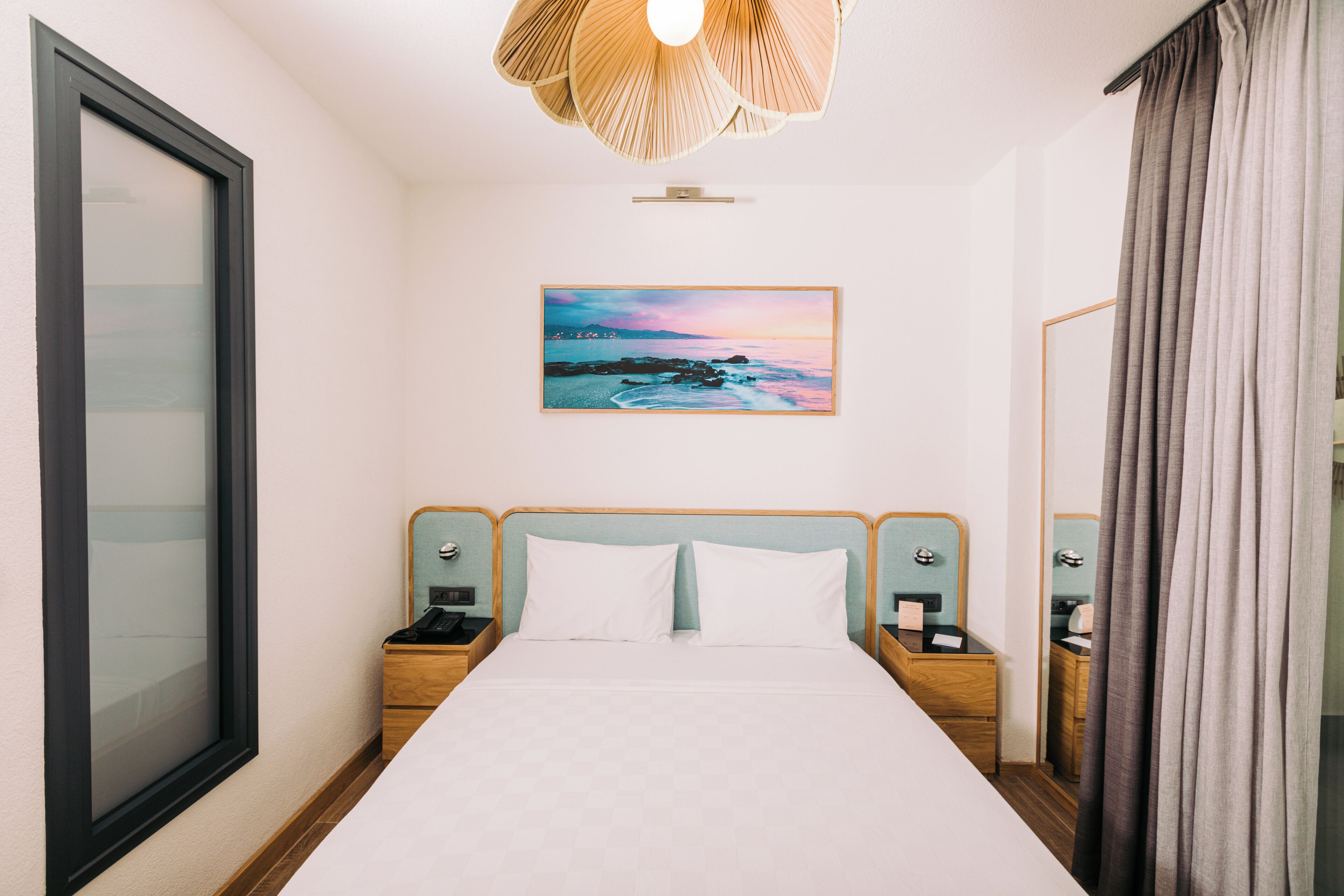A hotel room with a bed, nightstands, a painting of a beach scene, a window, and a curtain.