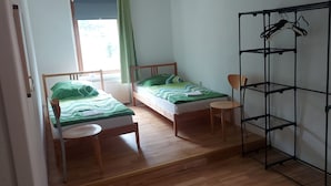 Una habitación pequeña y bien iluminada con dos camas de madera, un radiador, una silla y un estante metálico negro.