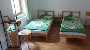 Una habitación con dos camas de madera, cada una con colcha verde y blanca estampada, una mesita de noche de madera con lámpara, y una silla.