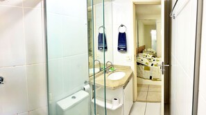 A bathroom with a glass shower enclosure, a toilet, a sink with a mirror, and a bed visible through an open door.