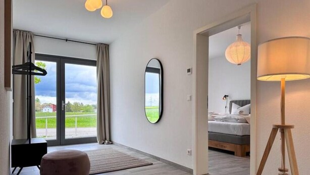 Ein modernes Hotelzimmer mit einer großen Glasschiebetür, die auf einen Balkon führt, einem Bett mit grauem Kopfteil, einem Holzboden und einem runden Spiegel an der Wand.