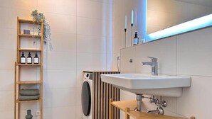 Ein modernes Badezimmer mit Holzregal, das Toilettenartikel, eine Waschmaschine und ein Waschbecken mit Spiegel beherbergt.