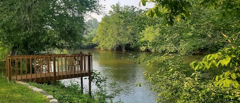 A wooden deck with chairs overlooking a calm river, surrounded by lush greenery.