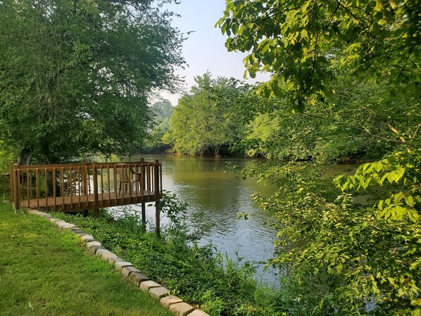 A wooden deck with chairs overlooking a calm river, surrounded by lush greenery.