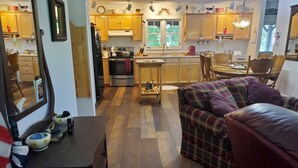 Een gezellige woonkamer met een geruite bank, een houten salontafel en een keuken op de achtergrond met houten kastjes en een fornuis.