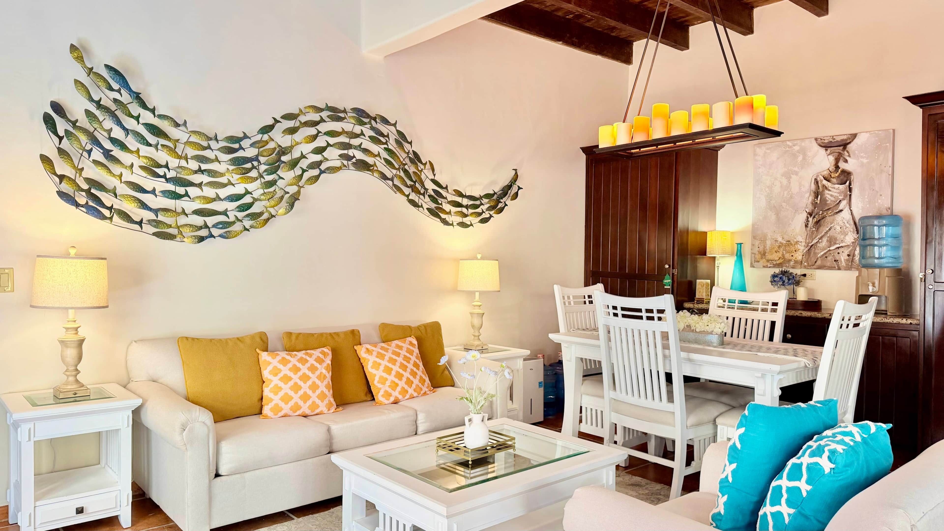 A living room with a white sofa, a glass coffee table, a dining area with white chairs, a wall art of fish, and a water dispenser.