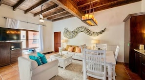 A living room with a sofa, coffee table, dining table, and chairs. There is a television, a decorative wall piece, and a chandelier. - Mission Gate 5B @ West Bay (West Bay)