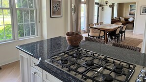 A modern kitchen with a gas stove, dining area with a wooden table and chairs, and large windows with a view of greenery. - Comfortable, bright home with heated pool/spa, large deck, and yard.   (Cape May)