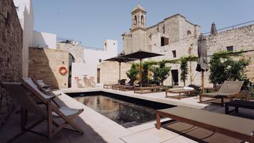 A courtyard with a swimming pool, lounge chairs, and a historic building with a bell tower.