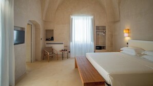 A bedroom with a large bed, a wooden bench, a desk with a chair, a television, a coat rack, and a window with curtains. - I Tre Bacili (Spongano)