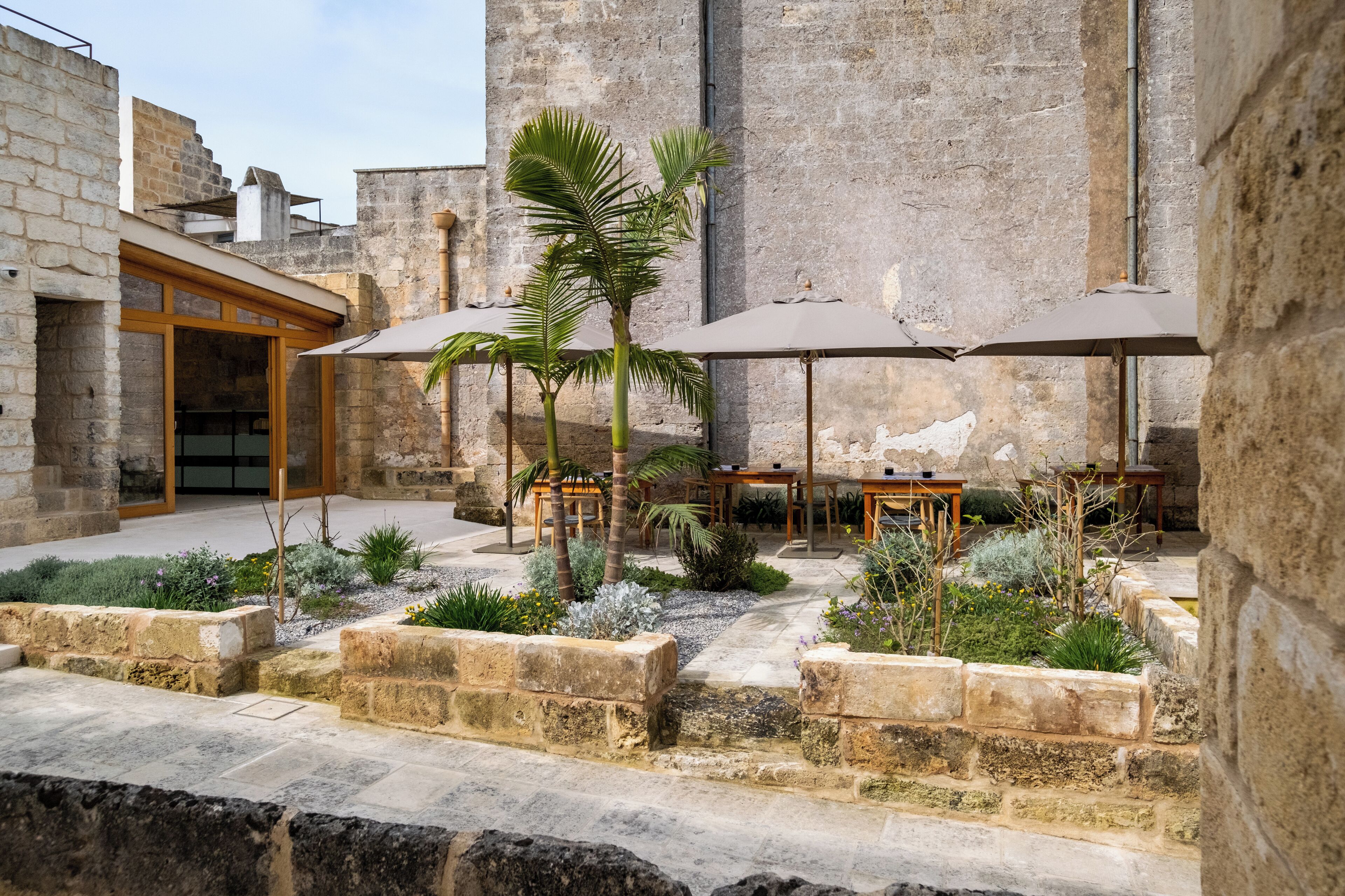 A courtyard with stone walls, outdoor seating, palm trees, and a building entrance.