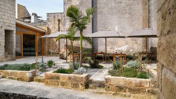A courtyard with stone walls, outdoor seating, palm trees, and a building entrance.