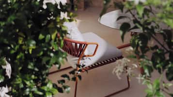 A wicker chair with a white cushion in an outdoor area with greenery and a concrete floor.