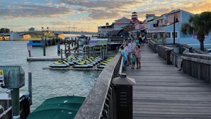 Un paseo marítimo frente al agua con barcos amarrados, un faro y diversas tiendas y restaurantes.