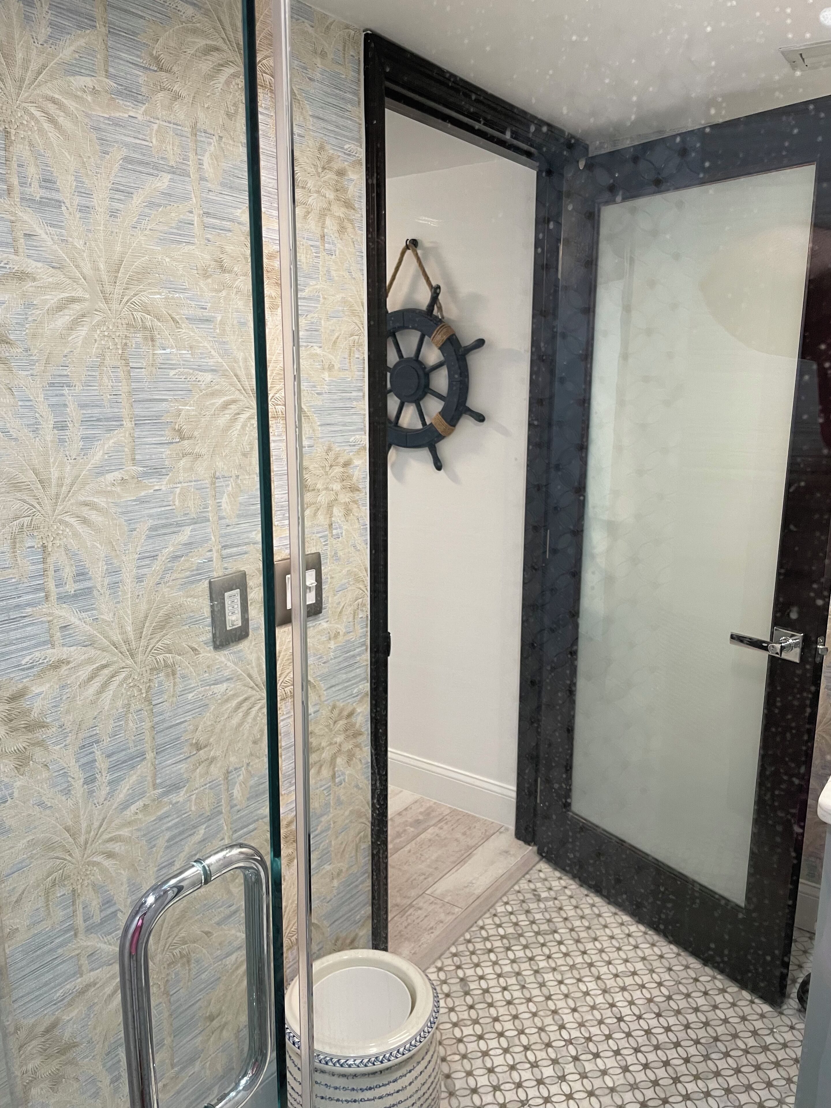 A modern bathroom with a glass shower enclosure, a ship's wheel decoration, and patterned tiles.
