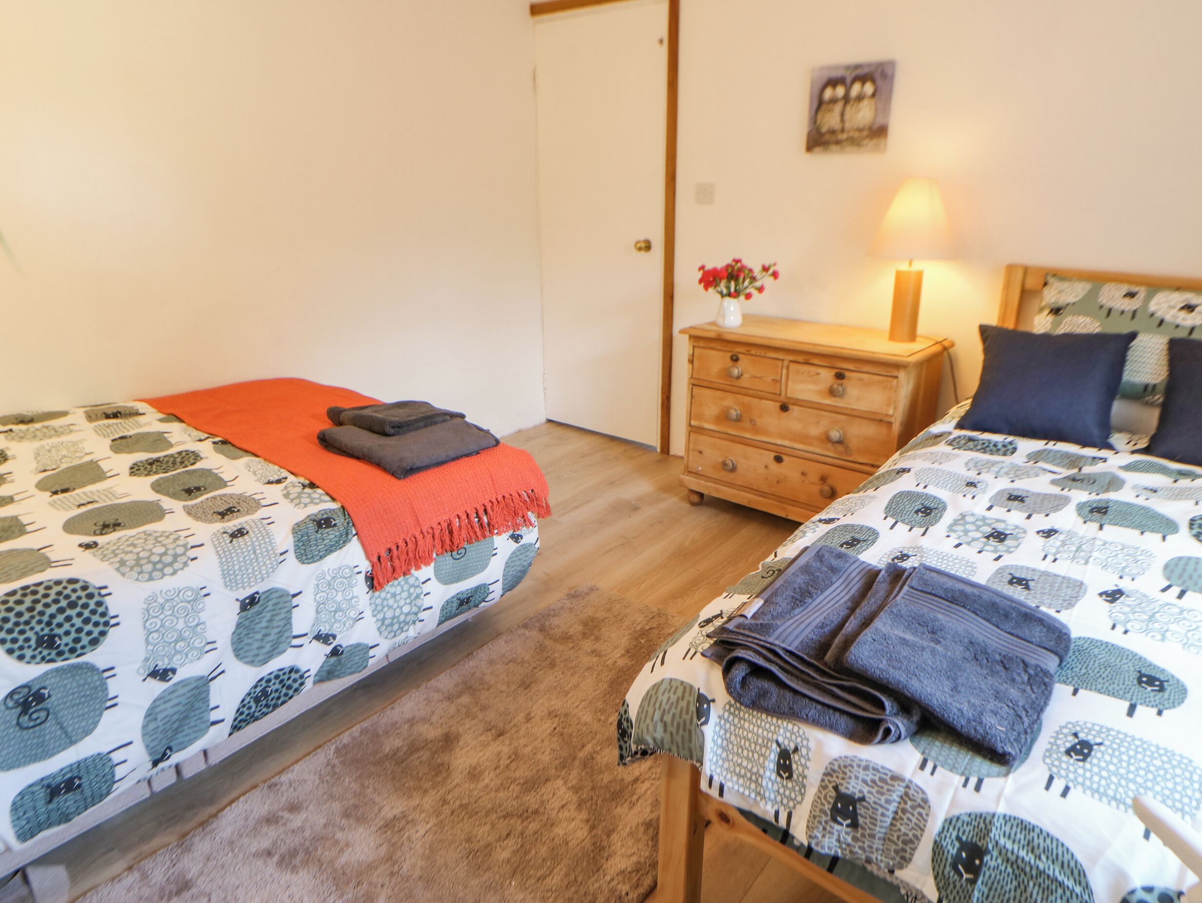 A bedroom with two beds, a wooden dresser, a nightstand with a lamp, and a wall with a framed picture.