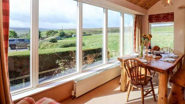 A dining area with a table set for two, large windows offering a view of the countryside, and a wooden ceiling. - Hepple Hill Cottage (Consett)