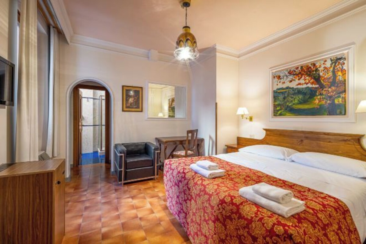 A hotel room with a bed, a desk, a chair, a television, and a painting on the wall.