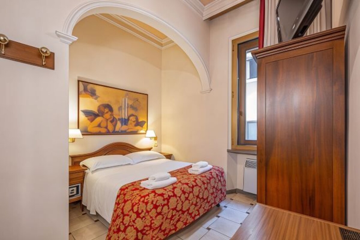 A hotel room with a bed, a wooden wardrobe, a painting on the wall, and a window.