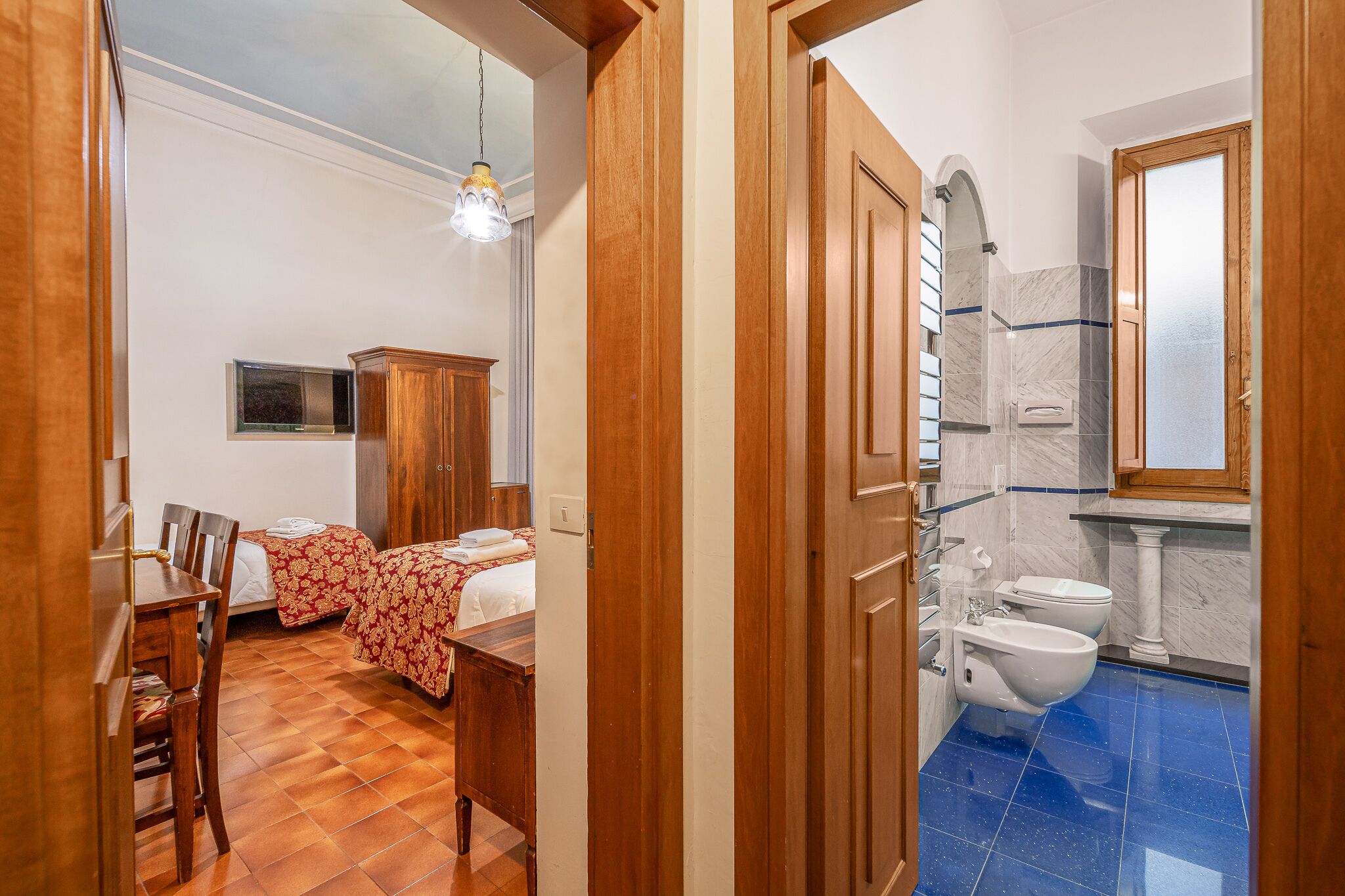 A hotel room with a wooden floor, two beds with floral bedding, a wooden wardrobe, a small table, and a bathroom with a blue-tiled floor, a white sink, and a toilet.