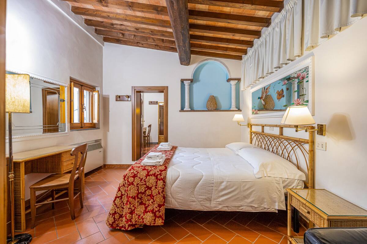A bedroom with a bed, a desk, and a chair. There is a window with curtains, a mirror, and a wall-mounted lamp.