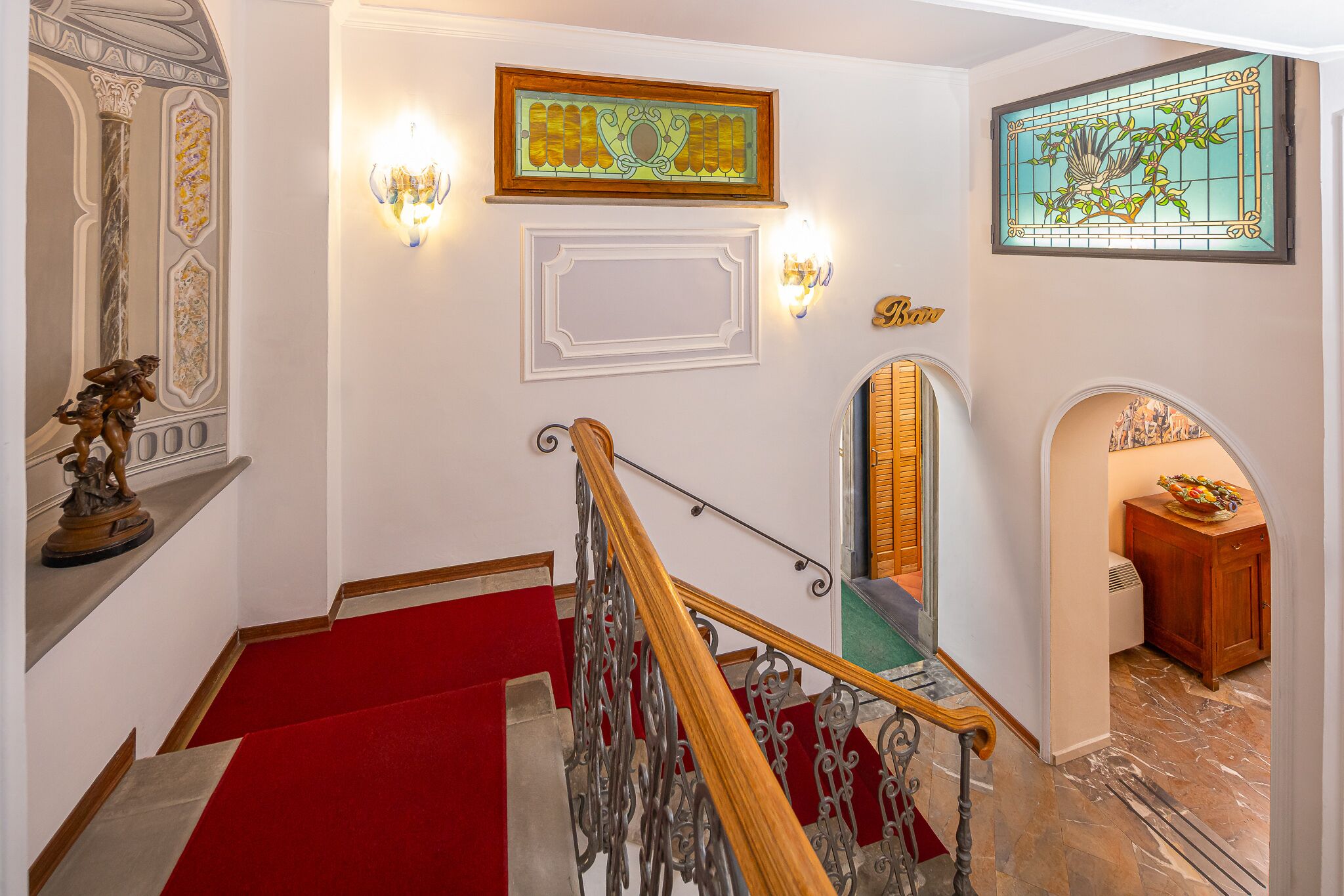 A grand staircase with red carpet, ornate wall decorations, and a chandelier.