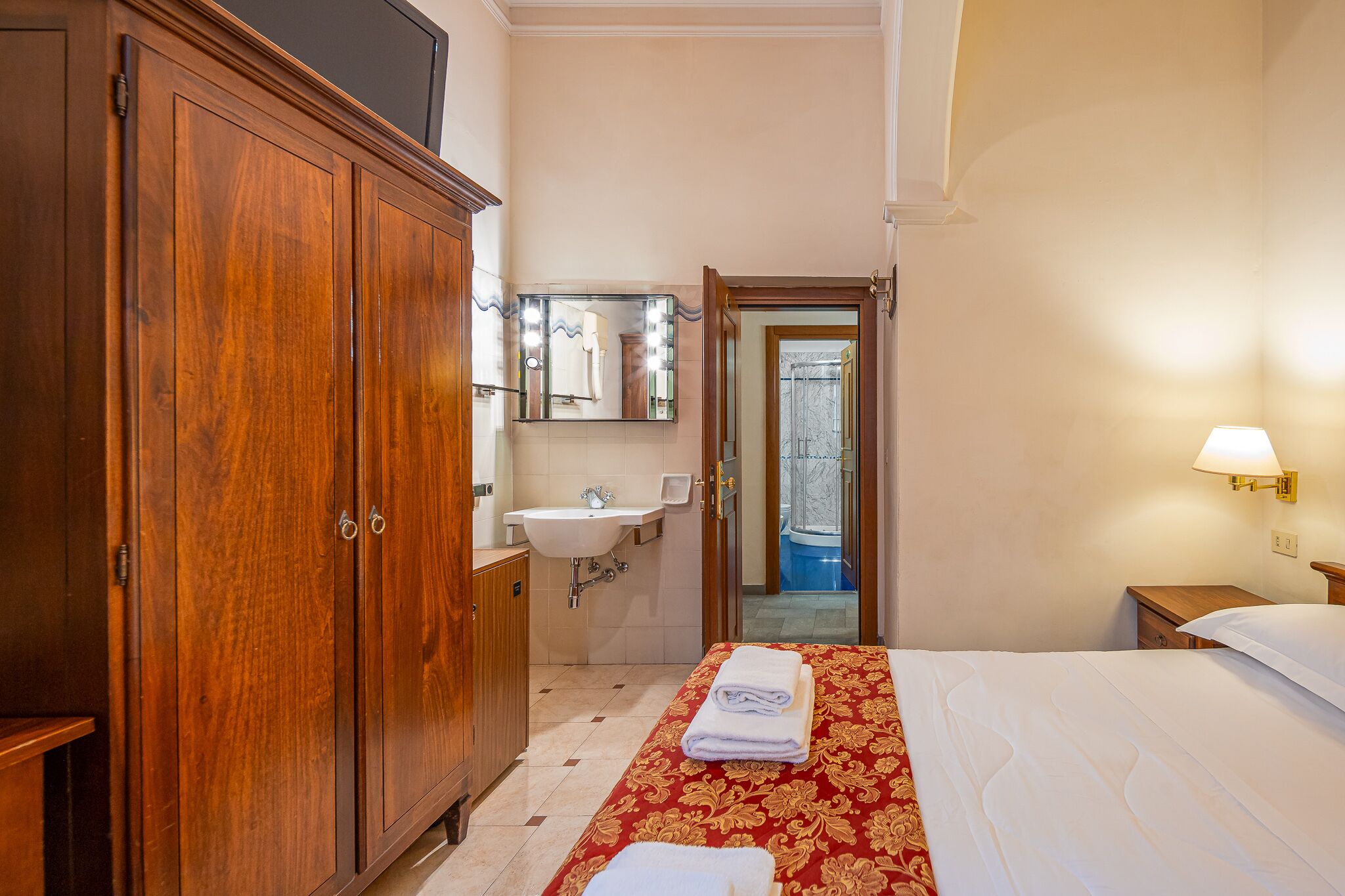 A hotel room with a wooden wardrobe, a sink, a mirror, a bed with a red and white bedspread, and a nightstand with a lamp.