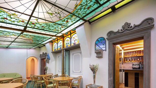 Una habitación con alfombra verde, muebles de mimbre, una zona de bar con botellas y ventanas de vitral.
