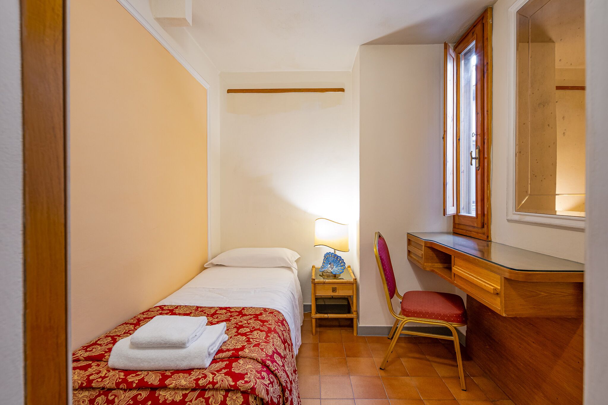 Una habitación pequeña y funcional con una cama individual, un escritorio, una silla y un espejo.