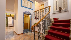 Una imponente escalera con peldaños alfombrados en rojo y barandales metálicos ornamentales, que conduce a una habitación con puerta de madera y ventana con marco azul.