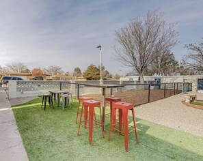 Relax in this outdoor seating area with a green lawn and a table for four, surrounded by a fence with a view of a building and a tree.