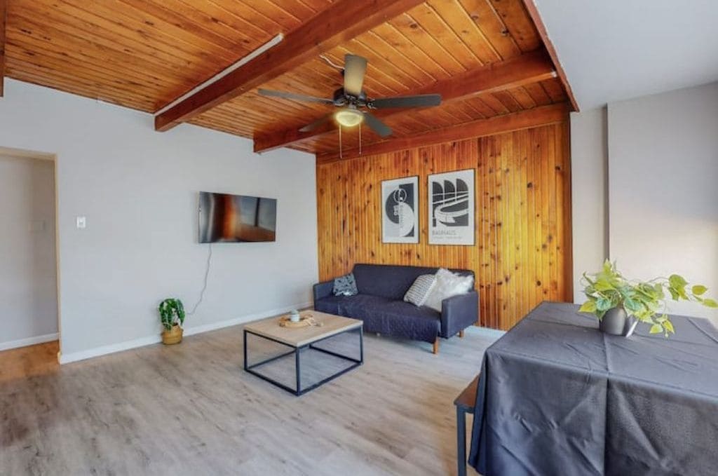 Ein modernes Wohnzimmer mit Holzboden, Deckenventilator, einem dunkelgrauen Sofa, einem Couchtisch und einem kleinen Tisch mit einer Pflanze.