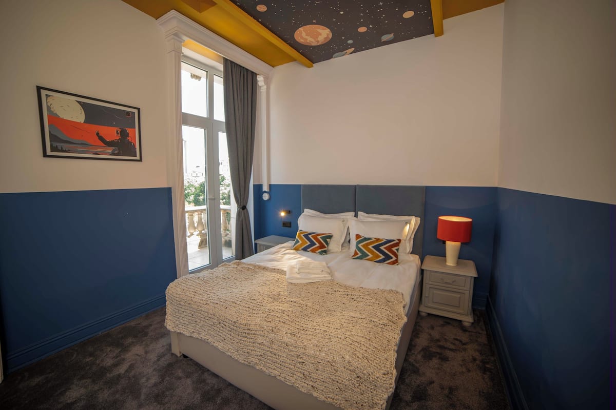 A bedroom with a bed, a nightstand, a lamp, a window with curtains, and a wall painting.