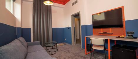 A room with a grey sofa, a desk with a TV, a small table, and a chair.