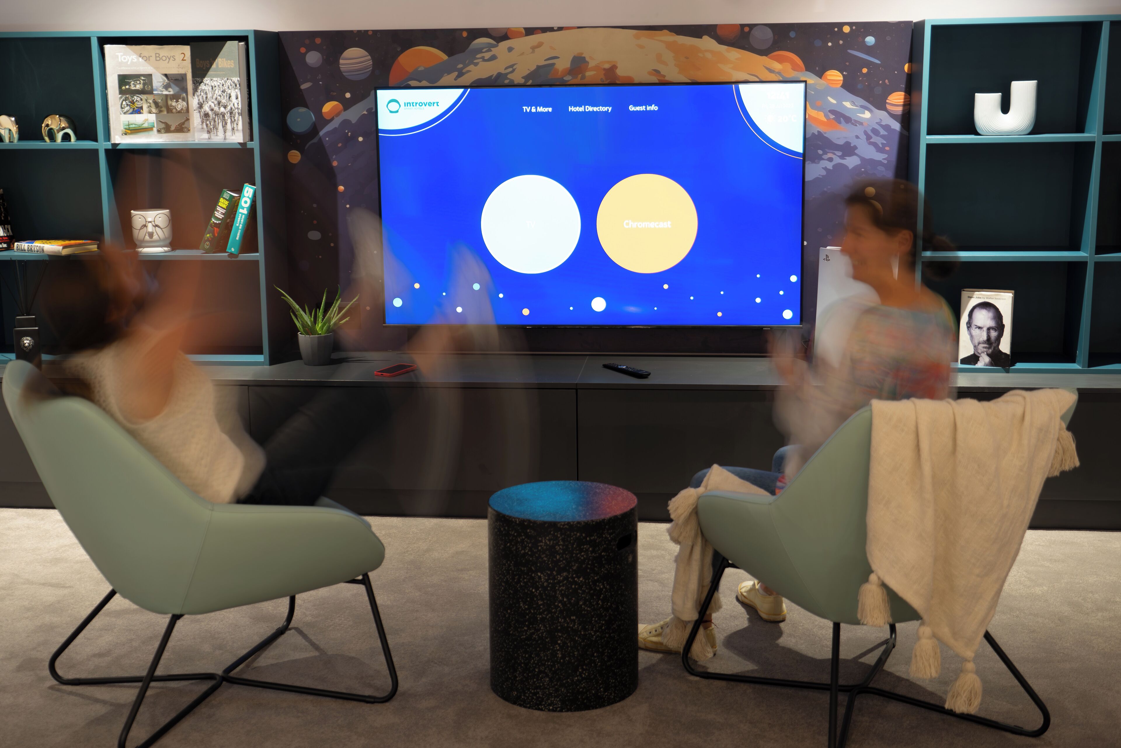 Un salon moderne doté d’un grand téléviseur à écran plat qui diffuse un papier peint sur le thème de l’espace. On y trouve deux fauteuils vert clair et une table basse ronde noire.