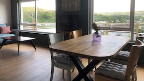 A modern living room with a wooden dining table, comfortable seating, large windows with a scenic view, and a flat-screen TV. - 2 bed apartment, waterfront views, 5 mins from shops & restaurants (Lochinver)
