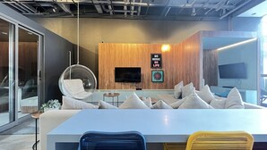 A modern office space with a large white desk, a hanging chair, and a sofa with multiple cushions. A flat-screen TV is mounted on the wall, and there is a framed picture with text. - MichiH - Perfeito Studio em Pinheiros (São Paulo)