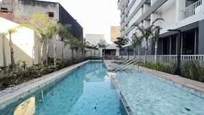 A rectangular swimming pool with clear blue water, surrounded by a tiled deck and a row of palm trees. - MichiH - Perfeito Studio em Pinheiros (São Paulo)