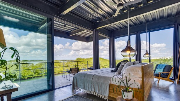 A modern bedroom with a large bed, a view of the outdoors, and a balcony with chairs. - Casa Balam: Private Chef Included - Waterfront Villa - 40ft Pool - Sunset Views (Placencia)