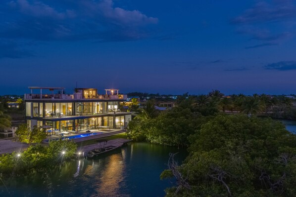 A modern multi-story house with a pool, located near a body of water surrounded by greenery. - Casa Balam: Luxury Waterfront Villa, Private chef incl, 40ft Pool & Rooftop Bar (Placencia)