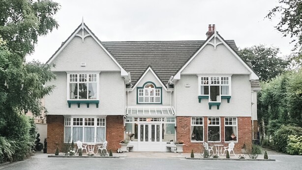 A two-story building with a symmetrical facade, featuring a central entrance with a glass door, flanked by brick and white walls, and a landscaped driveway with benches. - Herbert House (Killarney)
