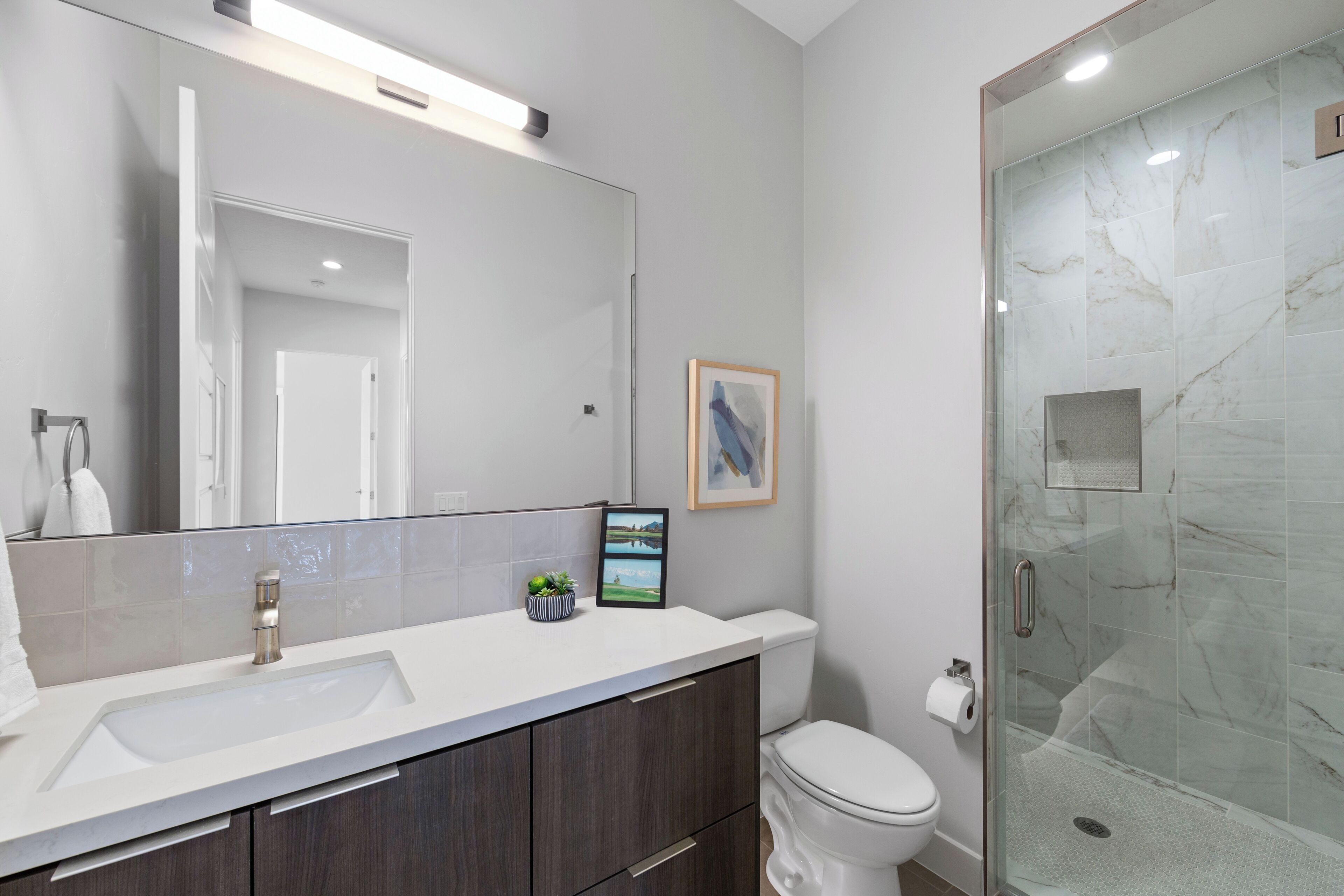 Un bany modern amb un lavabo blanc, armaris de fusta fosca, un vàter blanc i dutxa amb envà.