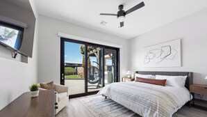 A modern bedroom with a large bed, a ceiling fan, a TV mounted on the wall, and a sliding glass door leading to an outdoor area.
