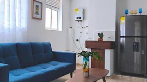 Una sala de estar con un sofá azul, una mesa de centro con una planta y una zona de cocina con un refrigerador y un dispensador de agua.