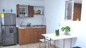 Cocina compacta con refrigerador, estufa, lavamanos y armarios de madera. Hay una mesa de comedor con un banco y una ventana arriba.
