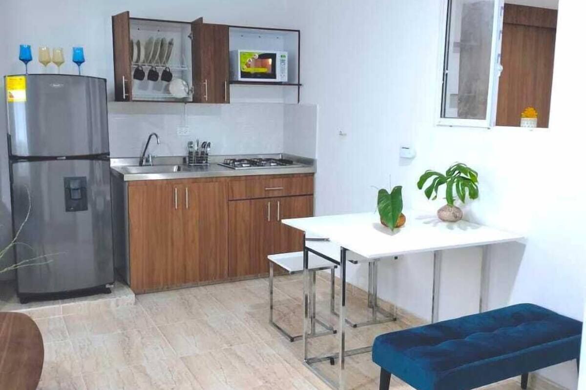 A compact kitchen with a refrigerator, stove, sink, and wooden cabinets. There is a dining table with a bench and a window above it.