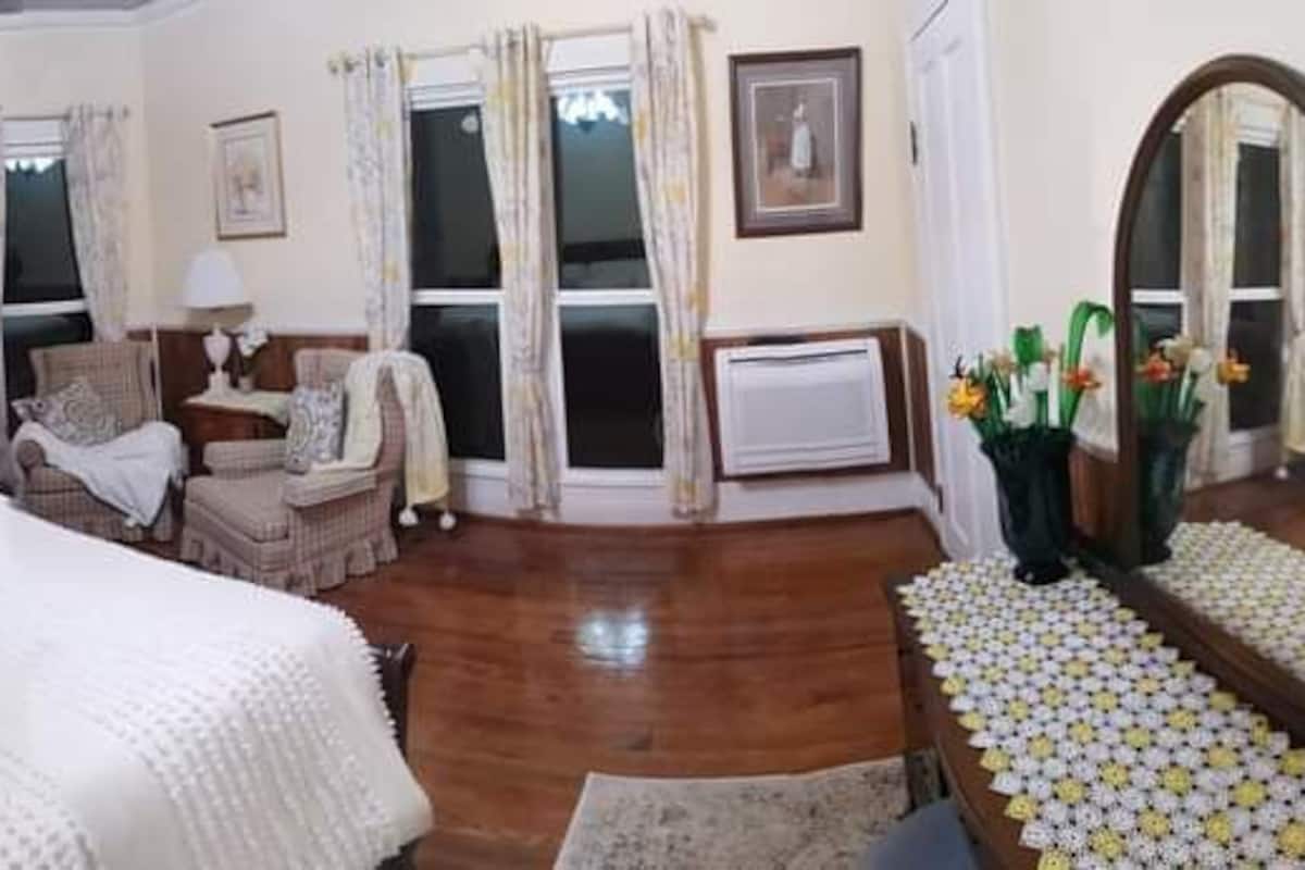 Une chambre dotée d’un grand lit, d’un coin salon avec un fauteuil et une table, et d’un miroir qui reflète la pièce.